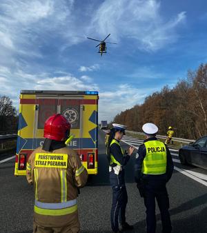 policjanci i strażacy stoją na autostradzie a nad nimi przelatuje helikopter