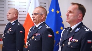 Trzech mężczyzn ubranych w mundur, za nimi flaga Polski i Unii Europejskiej.